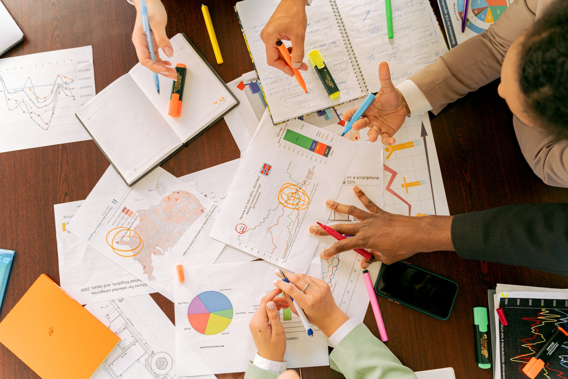 Picture showing charts, tables, notebooks and four individuals with pens on a table in a discussion.