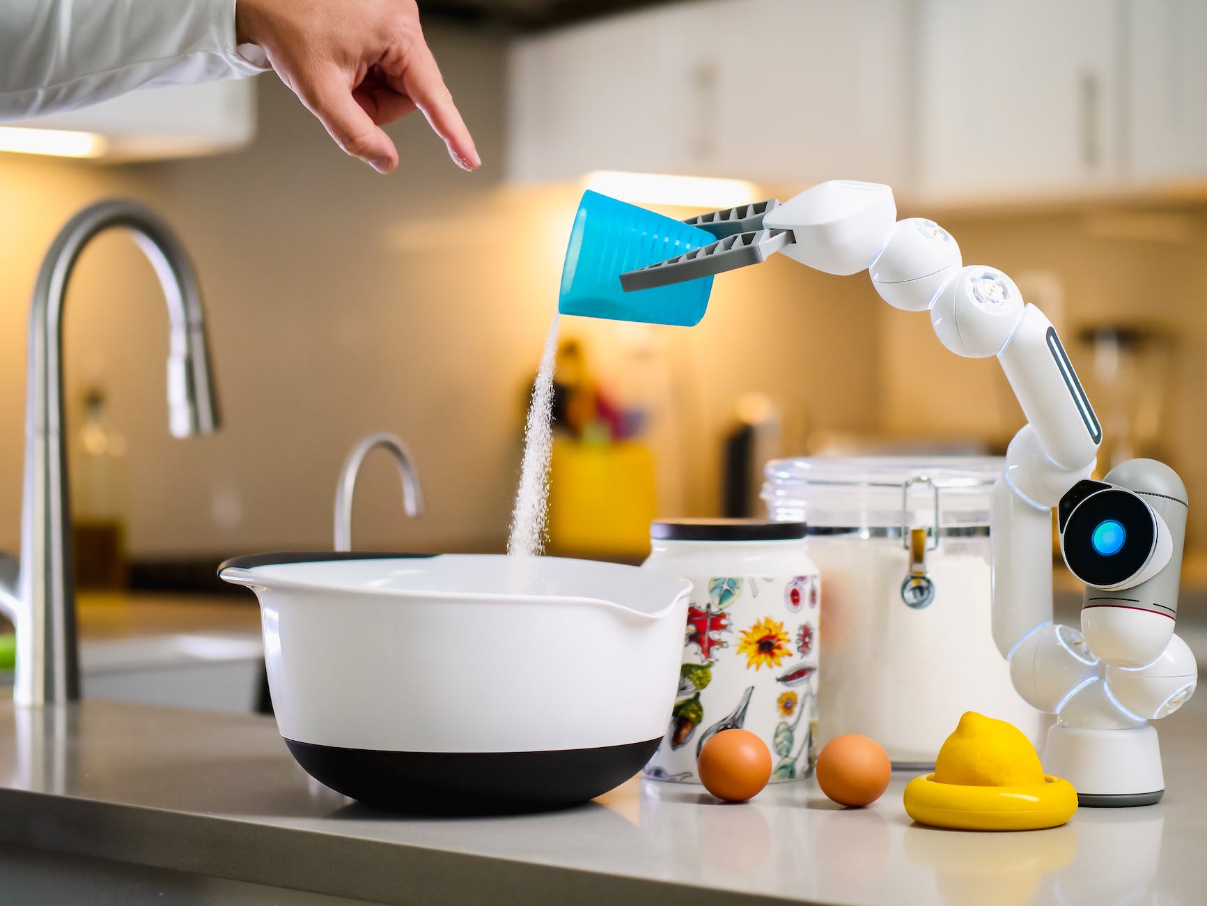 Picture showing a robotic holding a blue plastic cup pouring sugar into a bowl supervised by a human reflected by a hand.