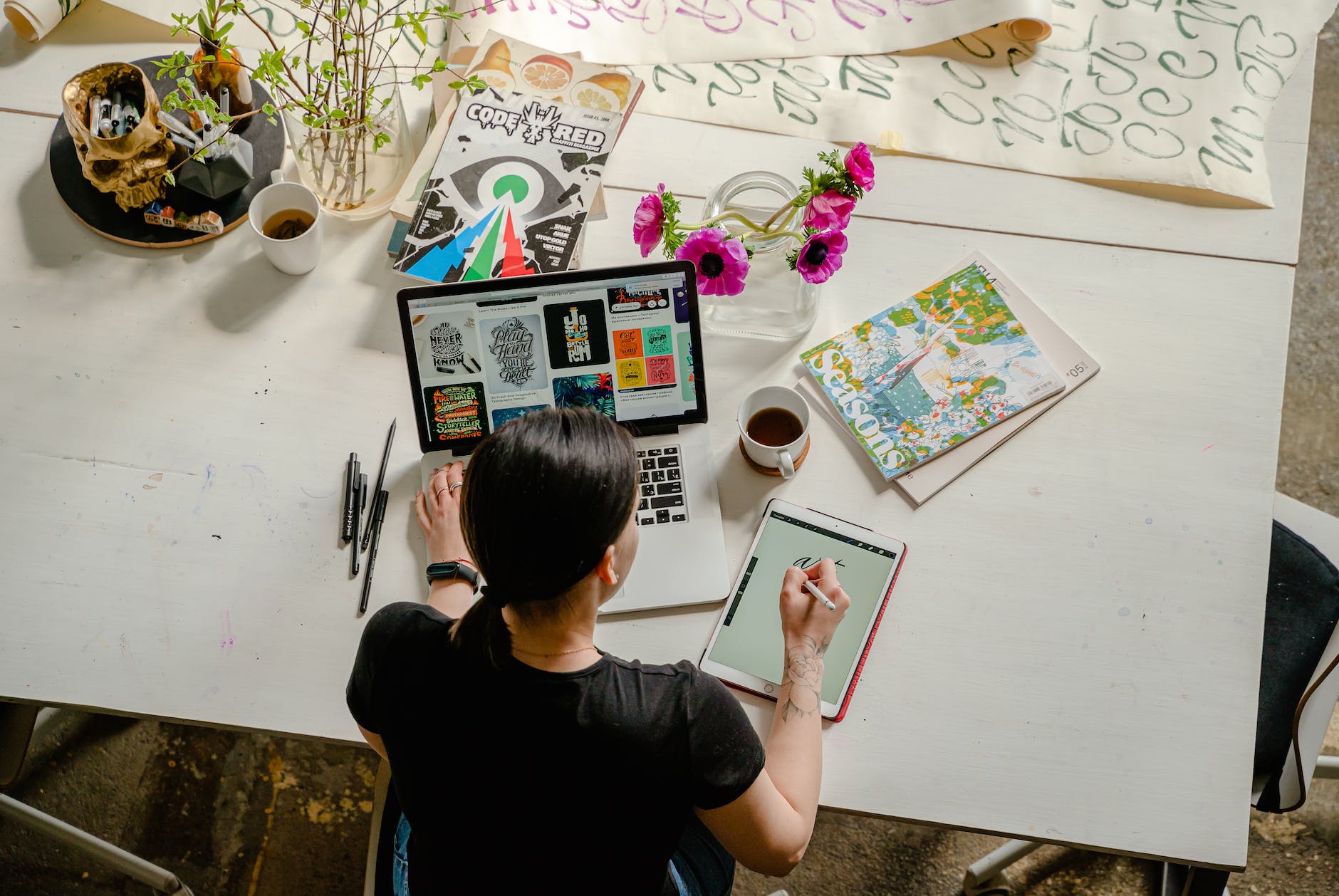 A girl working on a laptop and writing pad on a table with an array of things on the table, plants, coffee cup, magazines and art work.