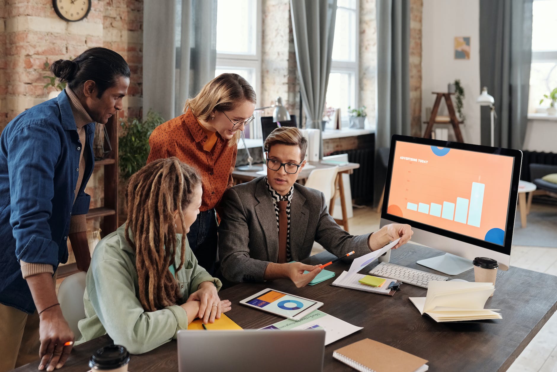 Four people in a discussion with a desktop and some books, files and stationery on a table.